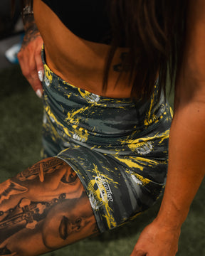 A woman in Born United Private Label Womens High-Waisted Seamless Front Compression Training Shorts | Unleash the Savage kneels on grass, showing tattooed arm and thigh, with her hand resting on her upper leg. She wears a black sports top.