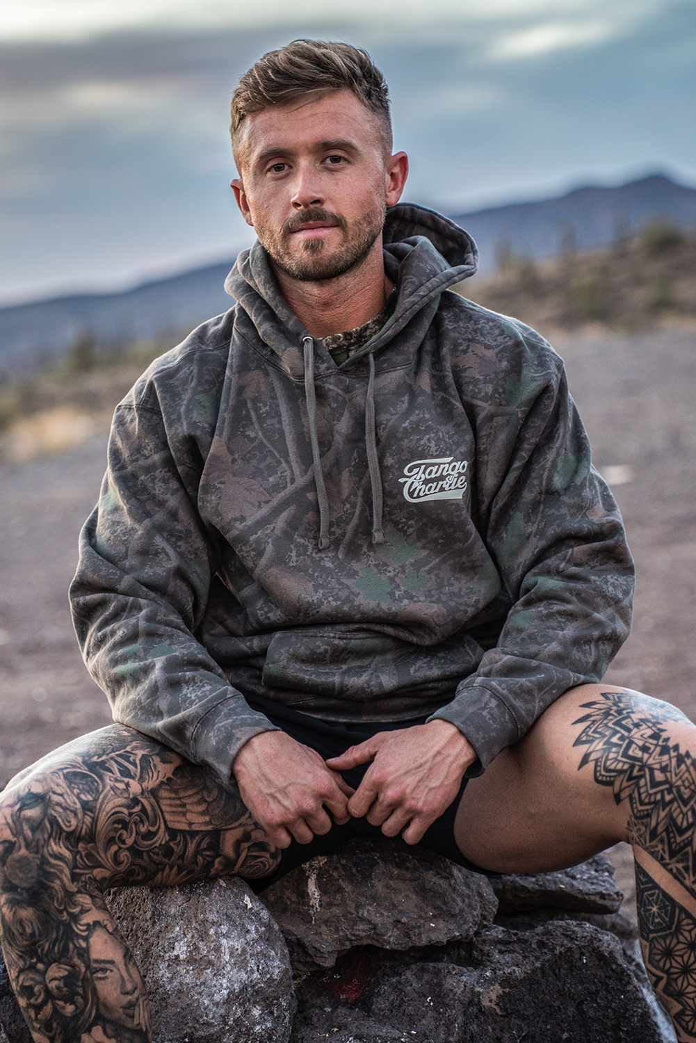 A tattooed man sits on rocky terrain outdoors, dressed in the Lift Heavy. Trust God. - Premium Darkwood Camo Hoodie by Tango Charlie Apparel. With a dramatic desert landscape and mountains behind him, he reflects the spirit of Christian Fitness—lifting heavy in both body and faith, inspired by Proverbs 3:5-6.