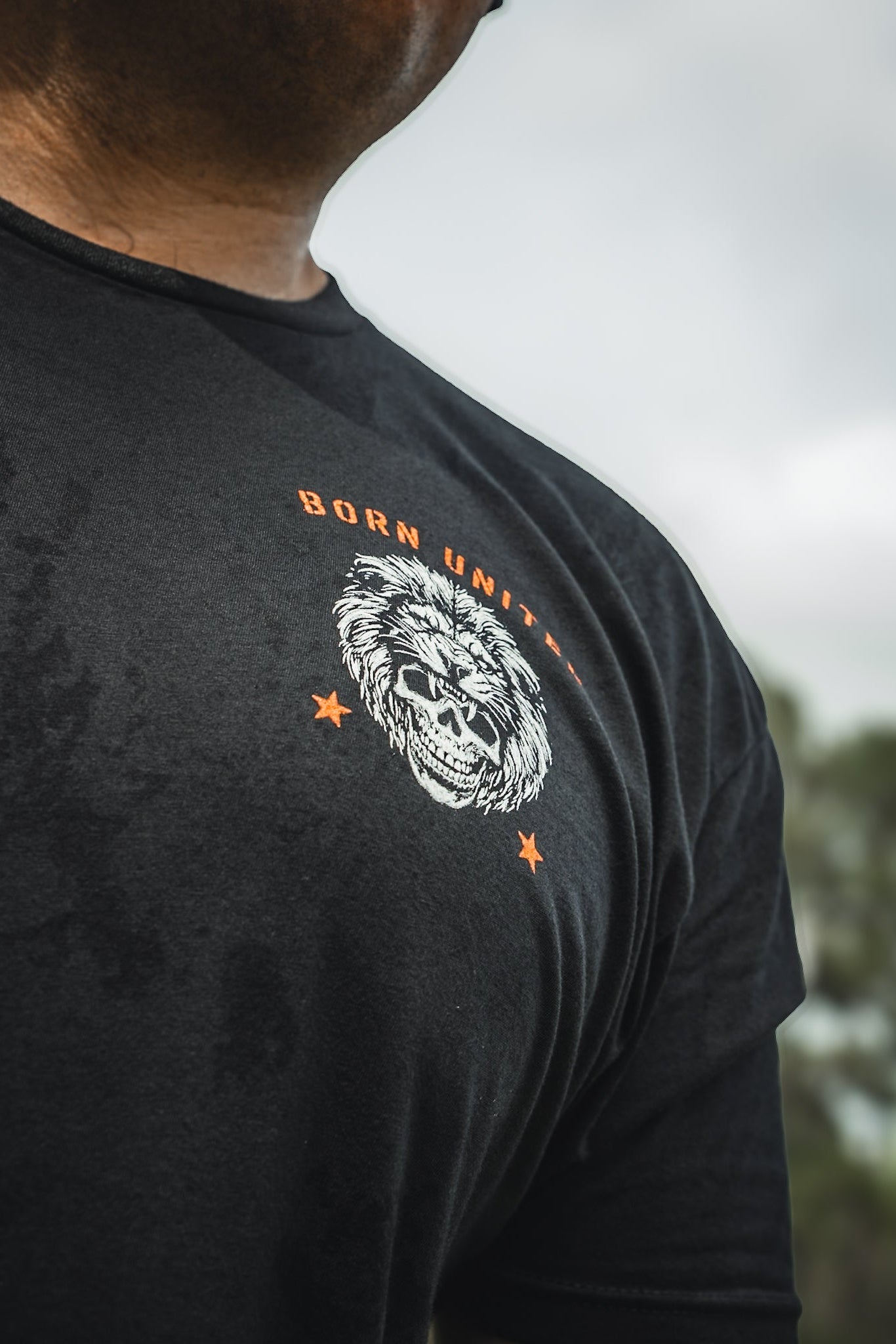 Someone wears a black Born United Private Label T-shirt named Waking Lions, with a fierce lions face graphic, the slogan WAKE THE LIONS, and two orange stars. The blurred background contains hints of greenery.