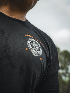Someone wears a black Born United Private Label T-shirt named Waking Lions, with a fierce lions face graphic, the slogan WAKE THE LIONS, and two orange stars. The blurred background contains hints of greenery.