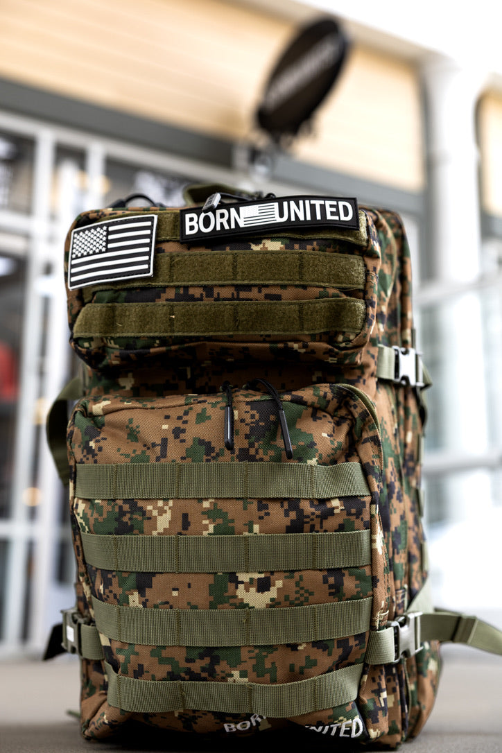 The Digi-Cam Tac-Pac - 45L by Born United Private Label features Marine Corps camo, an American flag patch with "BORN UNITED" on the top flap, and is pictured against a blurred outdoor background.