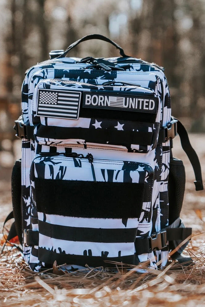 The Premium Tac-Pac Ultimate Bundle by Born United Private Label, featuring a black and white backpack with an American flag patch and BORN UNITED text, rests on dried pine needles amid a sunlit forest with blurred trees in the background.