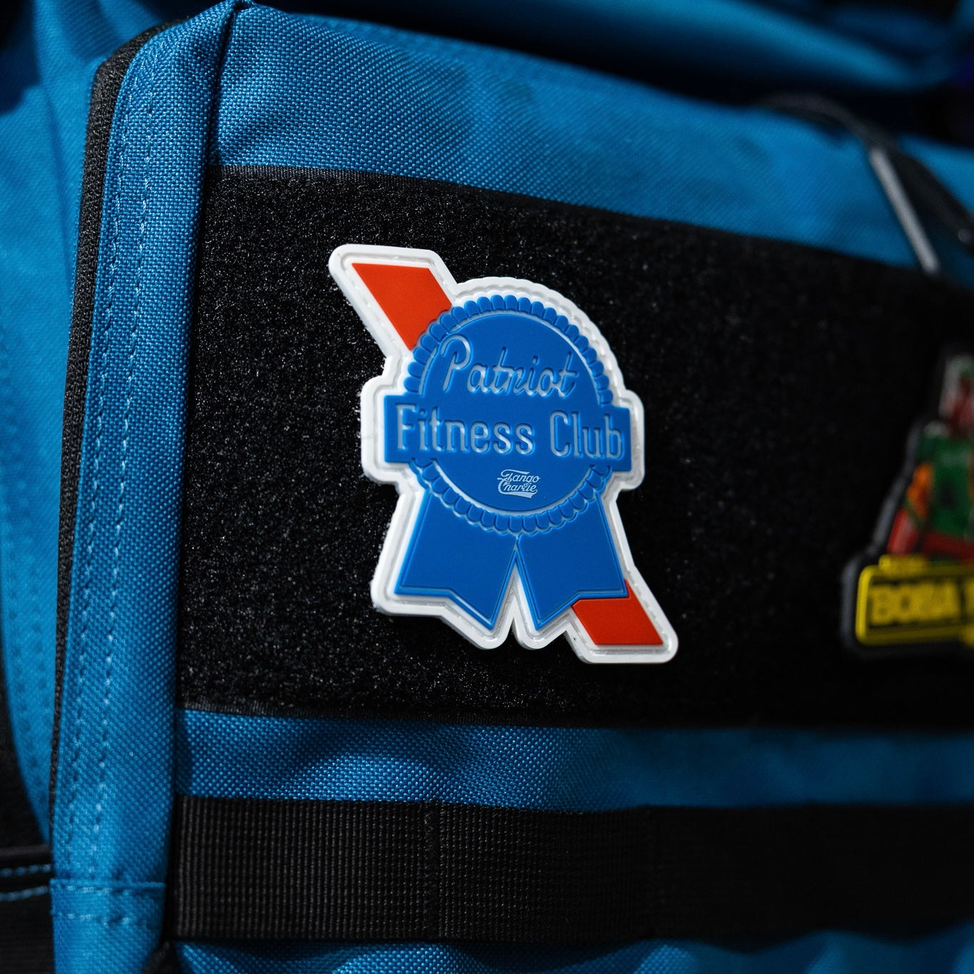 A Tango Charlie Apparel Patriots Fitness Club - Patch, shaped like a blue ribbon with red and white accents, is attached to the Velcro strip of a bright blue bag for a bold accessory.