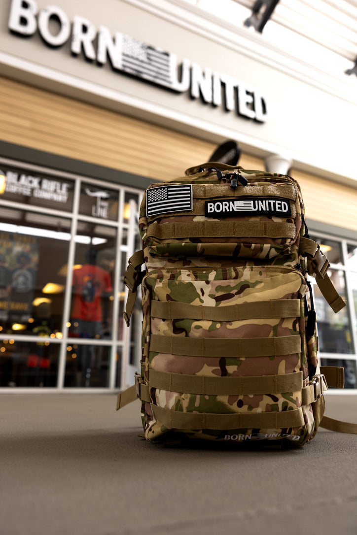 The Born United Private Label OCP Tac-Pac - 45L, featuring a camouflage design with an American flag patch and BORN UNITED label, stands outside a store showcasing matching signage, glass windows, and apparel inside.