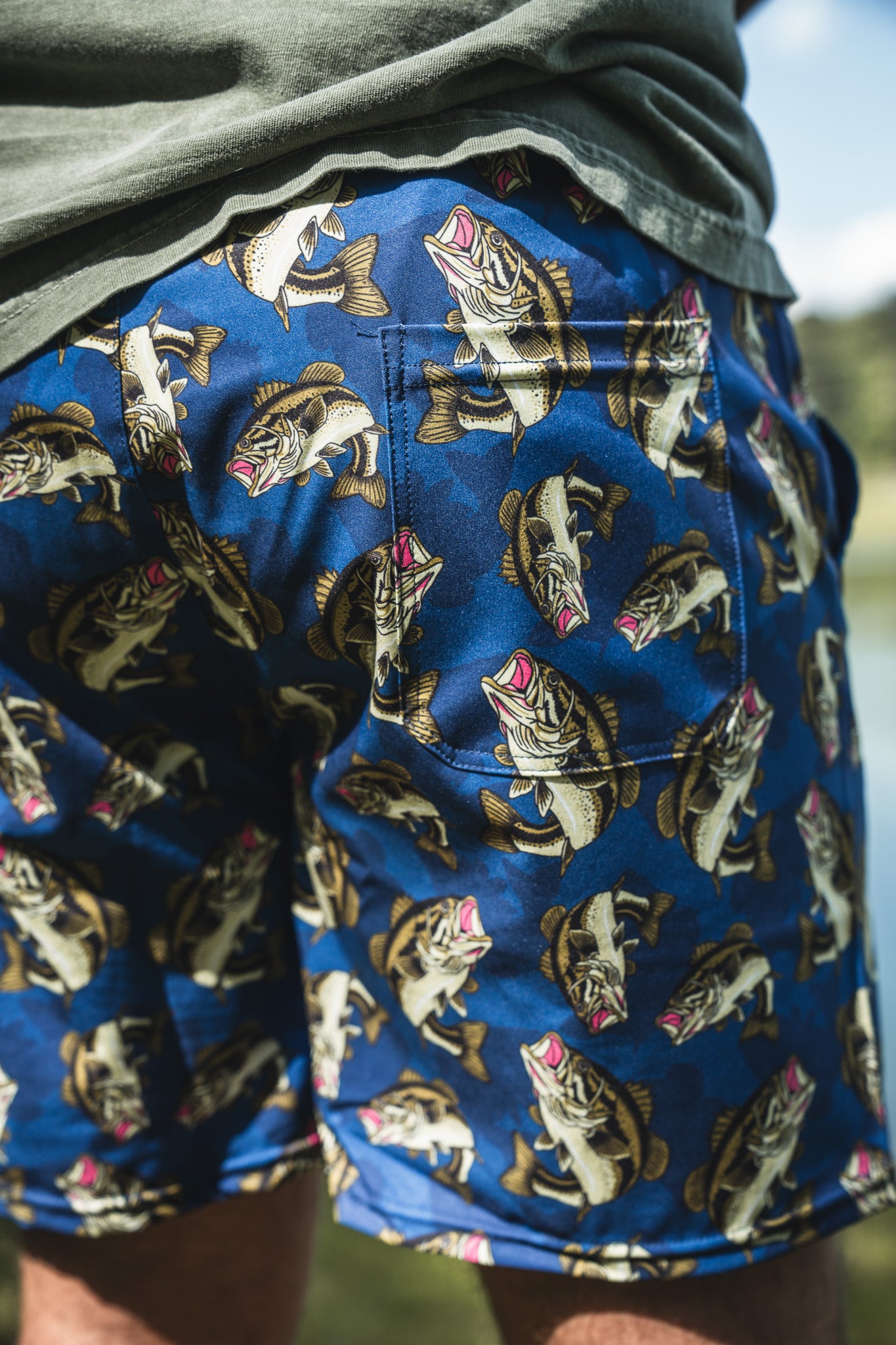 A close-up of a person wearing Mucker Bucket Men's Performance Athletic Shorts | 5.5"- Big Bass Daddy, featuring bold fish illustrations, standing outside on a sunny day.