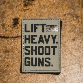 The LHSG - Patch by Tango Charlie Apparel is a 3-inch rectangular PVC patch on a rough brown surface, reading "LIFT HEAVY. SHOOT GUNS." in bold black letters with a small, partially obscured label at the top right corner.
