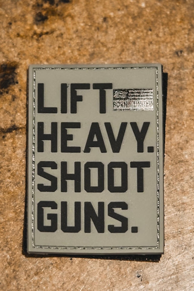 The LHSG - Patch by Tango Charlie Apparel is a 3-inch rectangular PVC patch on a rough brown surface, reading "LIFT HEAVY. SHOOT GUNS." in bold black letters with a small, partially obscured label at the top right corner.