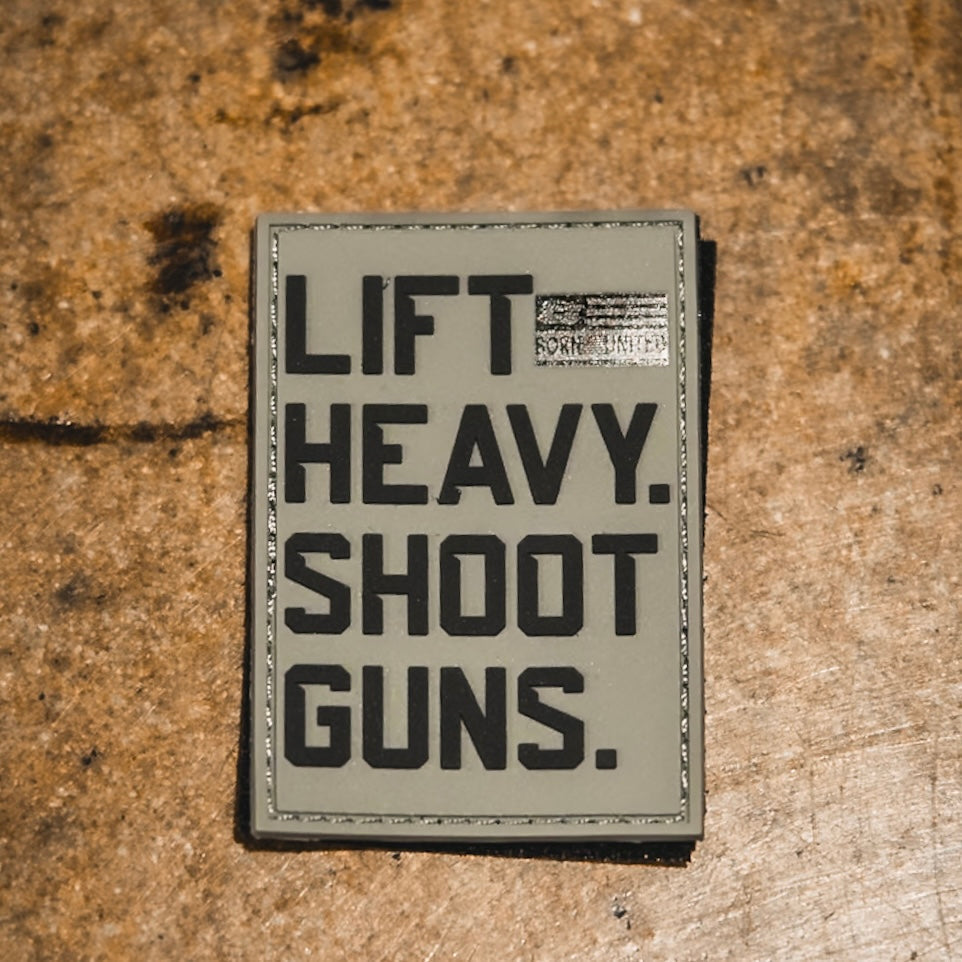 The LHSG - Patch by Tango Charlie Apparel is a 3-inch rectangular PVC patch on a rough brown surface, reading "LIFT HEAVY. SHOOT GUNS." in bold black letters with a small, partially obscured label at the top right corner.