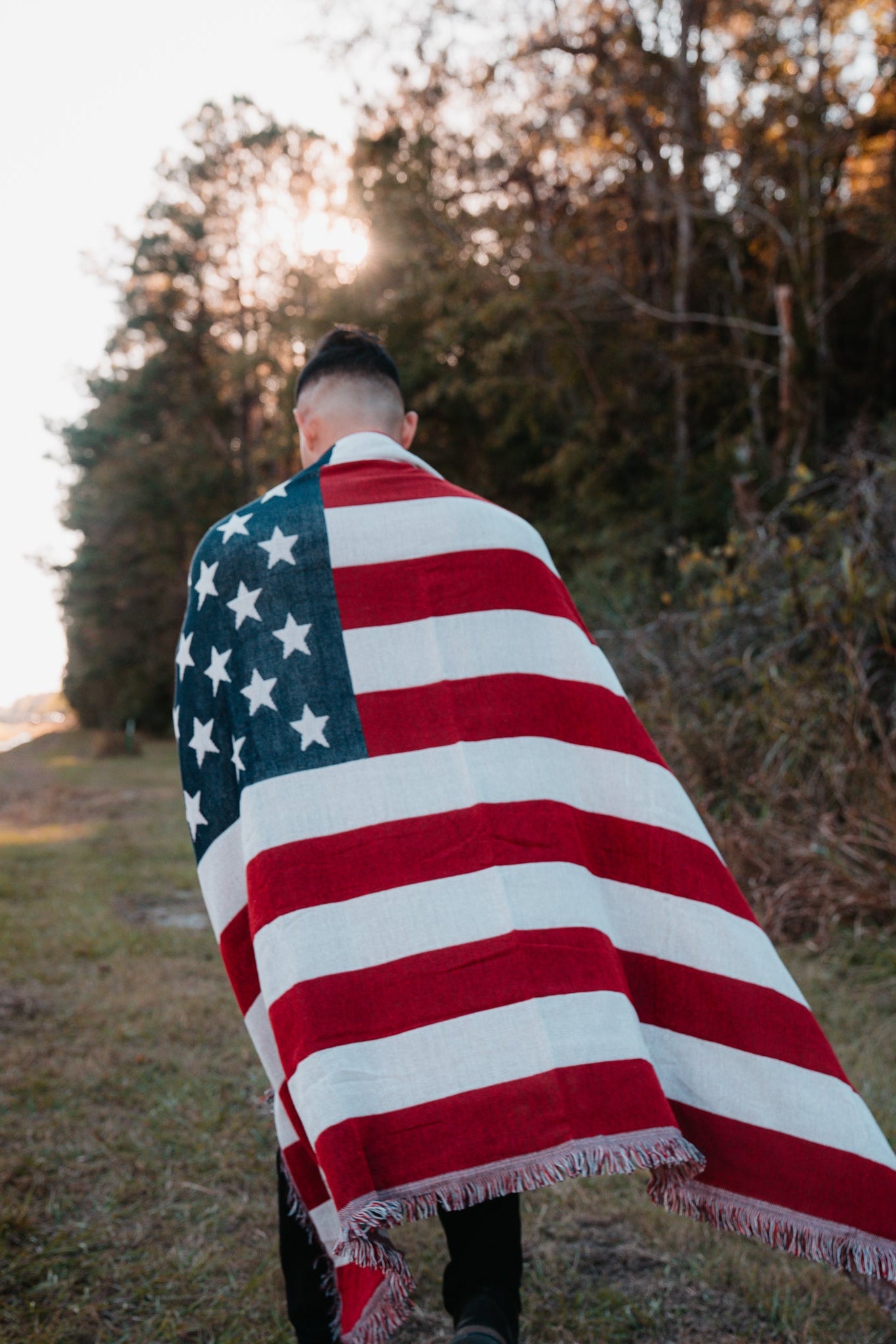 Wrapped in the American Flag Jacquard Blanket by Born United Private Label, a person strolls along a grassy path. The setting sun casts a warm light through the trees, creating a serene backdrop for the journey.