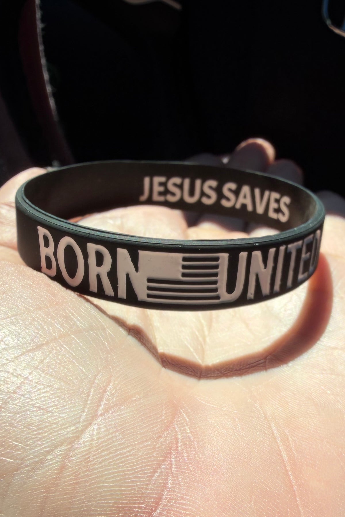 A hand displays the Born United Bracelet by Born United Private Label—a black silicone wristband with "BORN UNITED" and a flag on the outside, "JESUS SAVES" inside—illuminated by sunlight on the person’s palm.