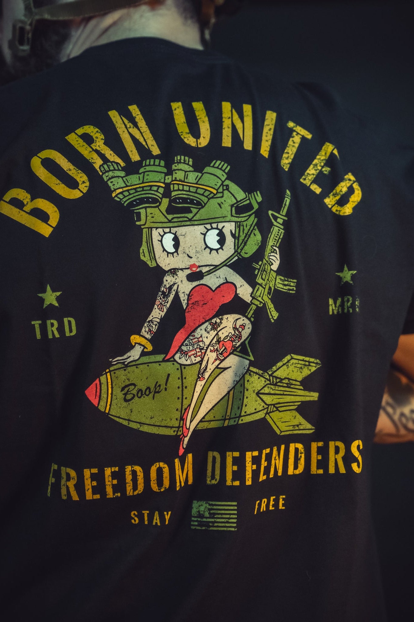 A person wearing the Betty Boop Tee by Born United Private Label, featuring a colorful cartoon pin-up girl sitting on a bomb with BORN UNITED above and FREEDOM DEFENDERS below, surrounded by stars and elements celebrating defenders.