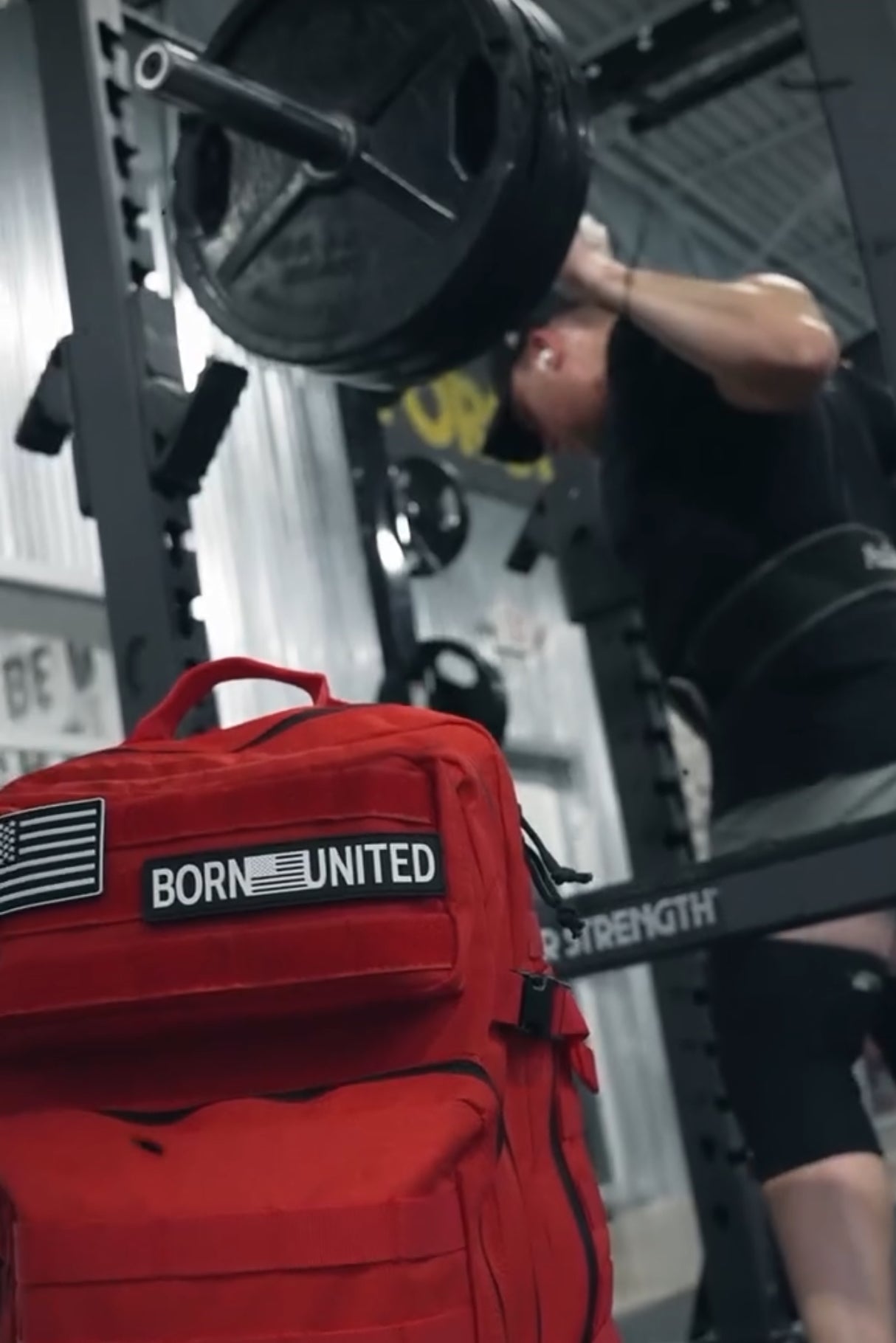 In a gym, someone squats with heavy weights on a barbell. In the foreground, a Revenge Red Tac-Pac - 45L by Born United Private Label with an American flag patch stands out against metal walls and strength equipment, embodying determination and resilience.