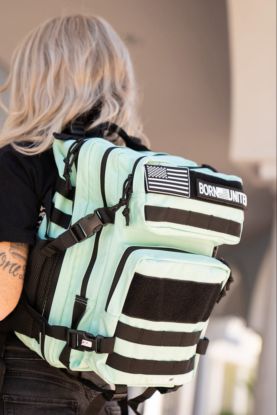 A person with long blonde hair and a black shirt is outdoors near a building, wearing the Mint Condition Tac-Pac - 25L by Born United Private Label, featuring light blue and black stripes plus an American flag patch.