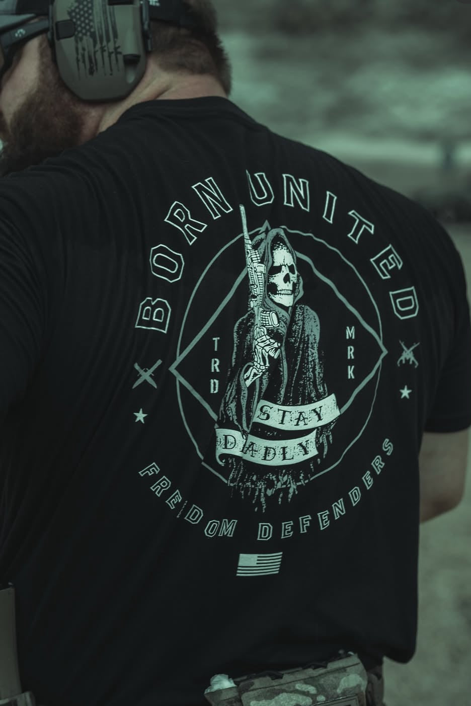 Wearing the Stay Deadly Tee - Black by Born United Private Label, featuring a Grim Reaper with a rifle and the text BORN UNITED, STAY DEADLY, and FREEDOM DEFENDERS, paired with headphones and camo pants for a stylish, fierce look.