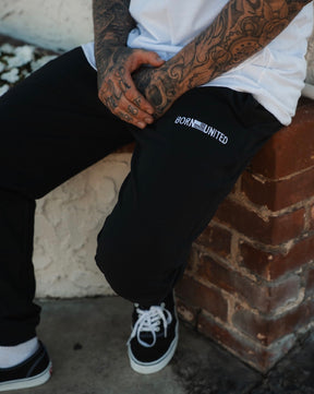 A person with tattooed hands sits on a brick ledge, wearing a white shirt, black-and-white sneakers, and Men’s Classic Premium Joggers | Stealth by Born United Private Label. Their hands are clasped over their lap.