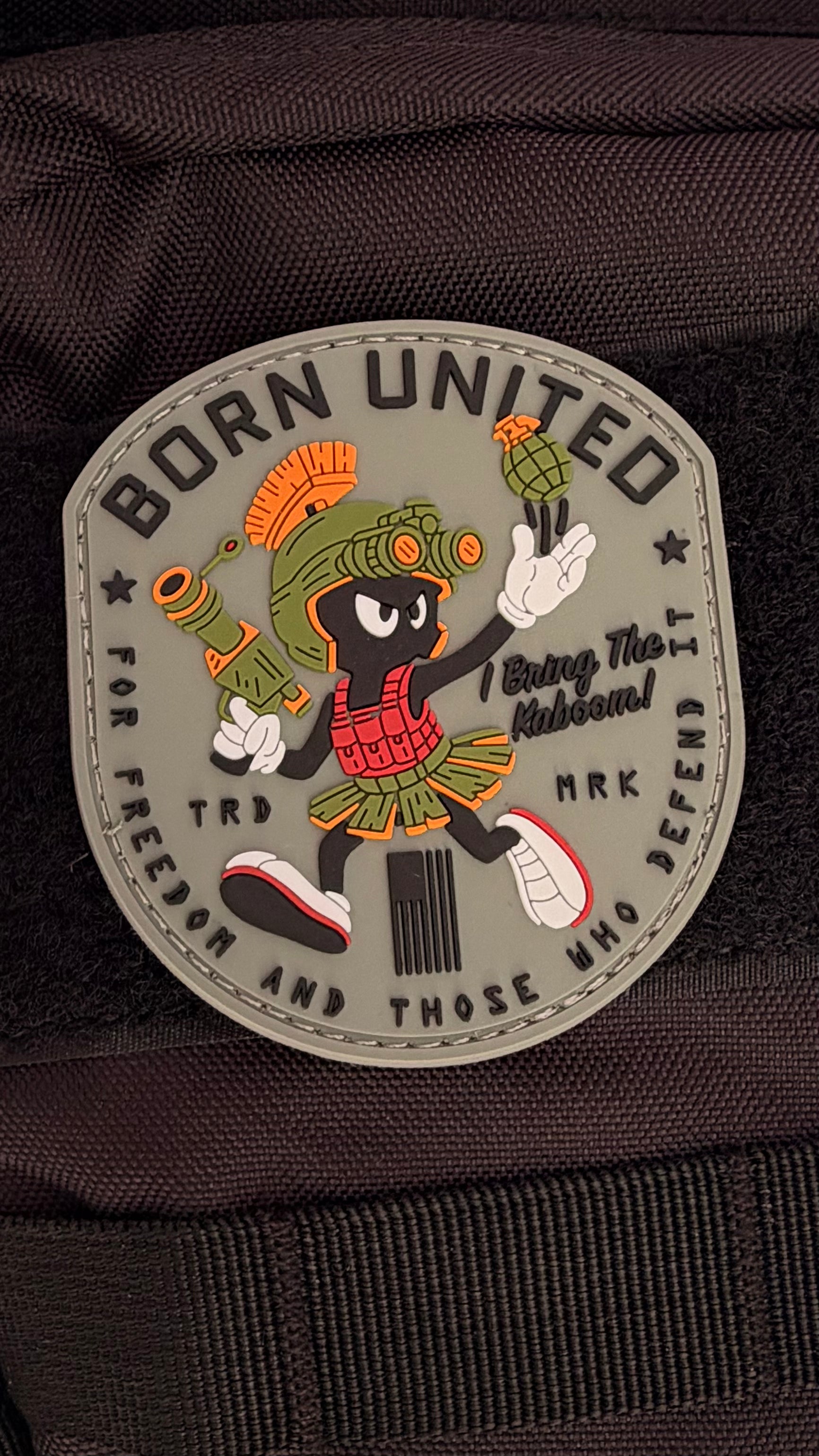 The Kaboom! PVC Patch by Born United Private Label features a cartoon Martian in Roman armor with a grenade and futuristic gun, plus "BORN UNITED" and "For freedom and those who defend it" text on a dark background.