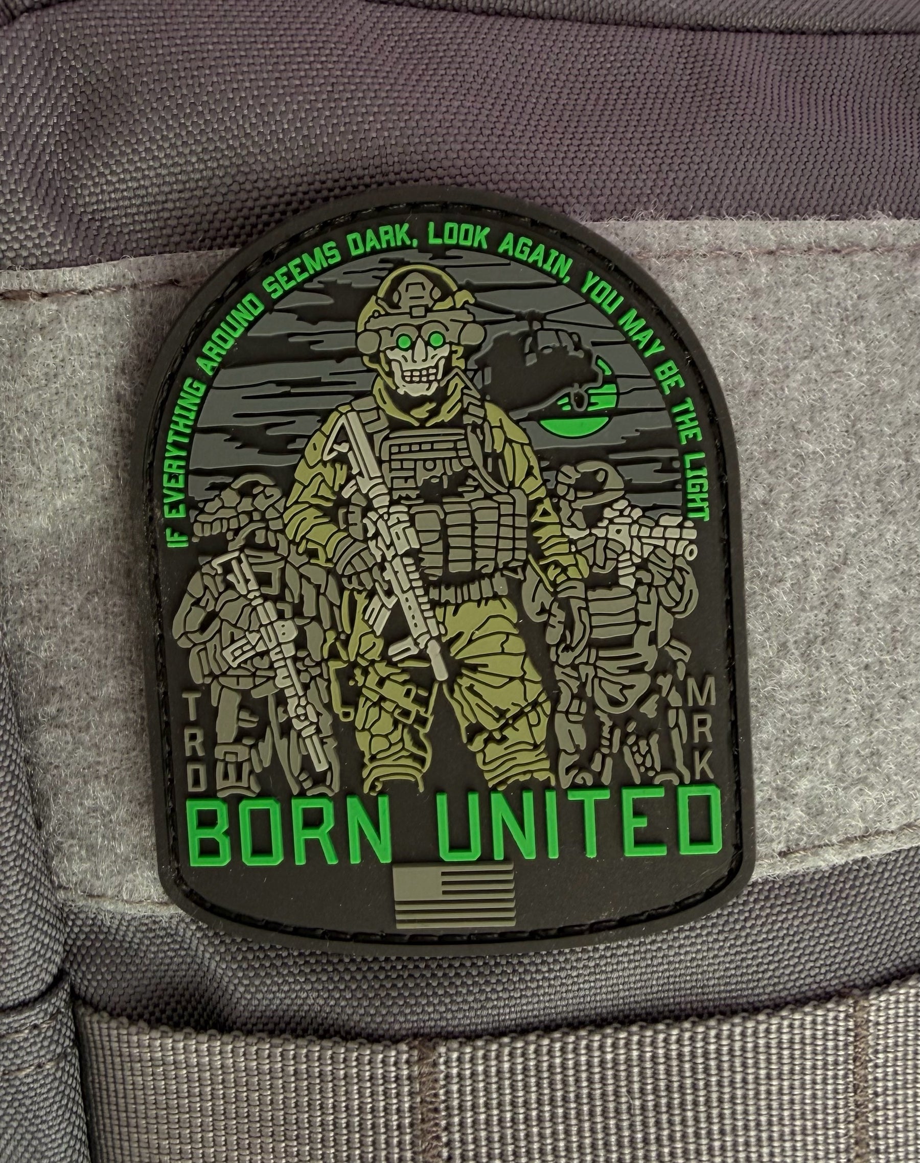 The Light in the Darkness PVC Patch by Born United Private Label features three armed soldiers with night vision goggles, a helicopter backdrop, and green text: “If everything around seems dark, look again, you may be the light. Born United.”.