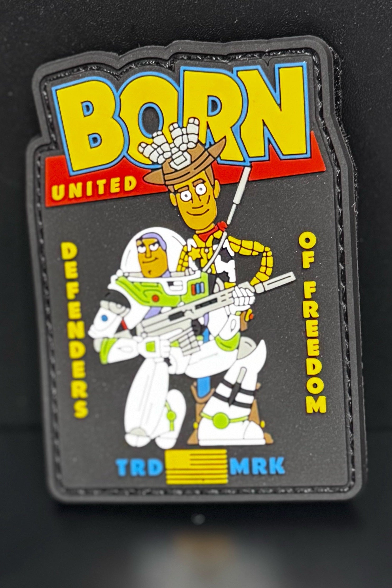 The Buzz N Woody PVC Patch by Born United Private Label features Buzz holding a rifle and Woody in a helmet with bullets. The patch reads BORN UNITED DEFENDERS OF FREEDOM TRD MRK and includes a yellow-striped design at the bottom.