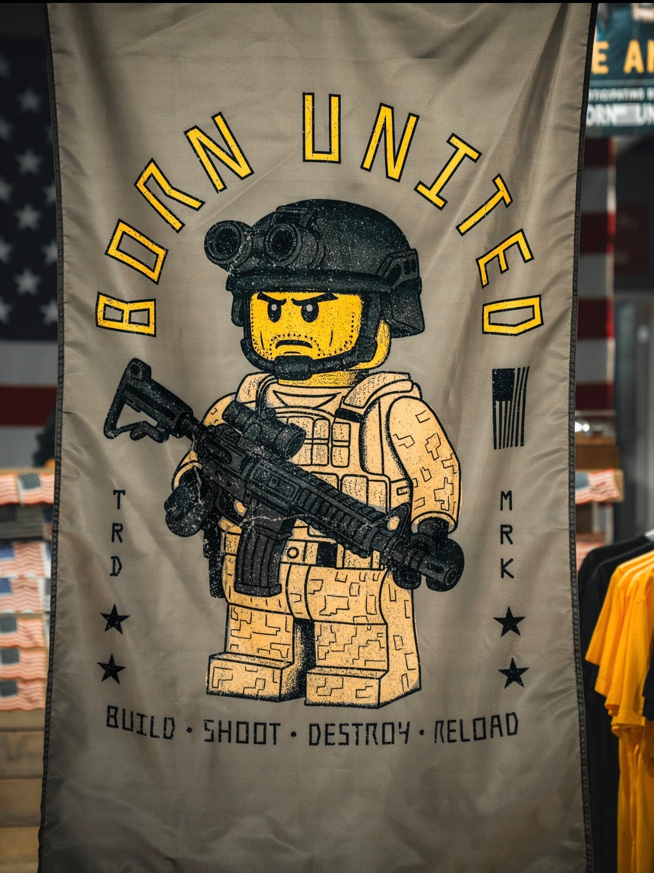 The Brick Operator Flag by Born United Private Label features a LEGO-style soldier in desert camo with a rifle, set against an American flag and surrounded by “BORN UNITED,” “BUILD SHOOT DESTROY RELOAD,” and stars and stripes.
