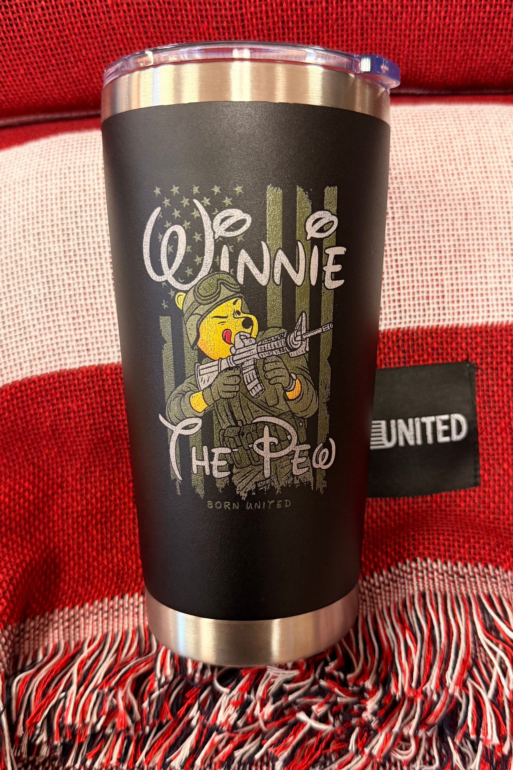 The Born United Private Label Winnie the Pew Tumbler - 20oz features a cartoon bear in military gear with “Winnie the Pew” text and sits on a red and white fringed blanket with a black "UNITED" tag.