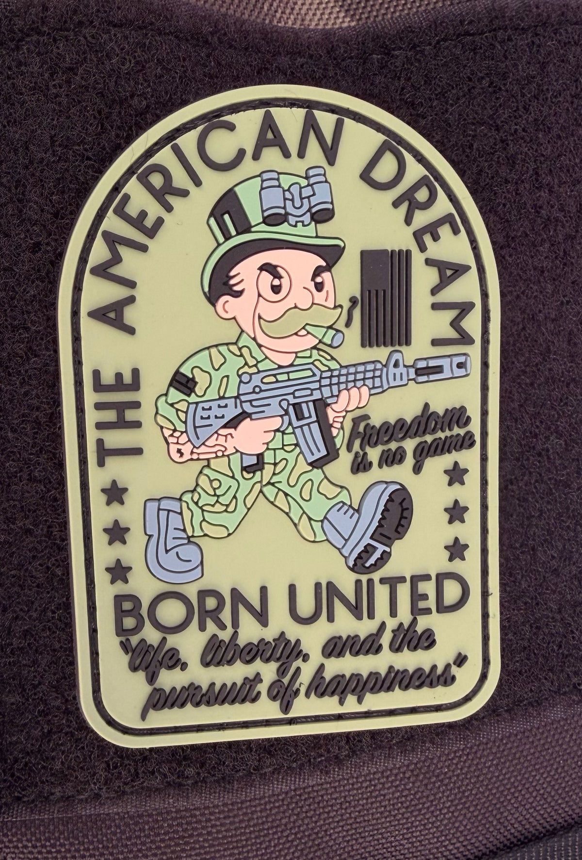 The American Dream PVC Patch by Born United features a cartoon soldier marching with rifle and text: "THE AMERICAN DREAM. Freedom is no game. BORN UNITED. Life, liberty, and the pursuit of happiness.