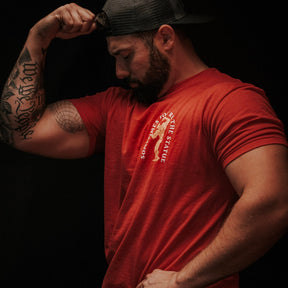 A man wearing Tango Charlie Apparel's "St. Arnold The Stoic" shirt and a backwards cap flexes his tattooed right arm, showing off his bicep against a dark background as he looks down at it.