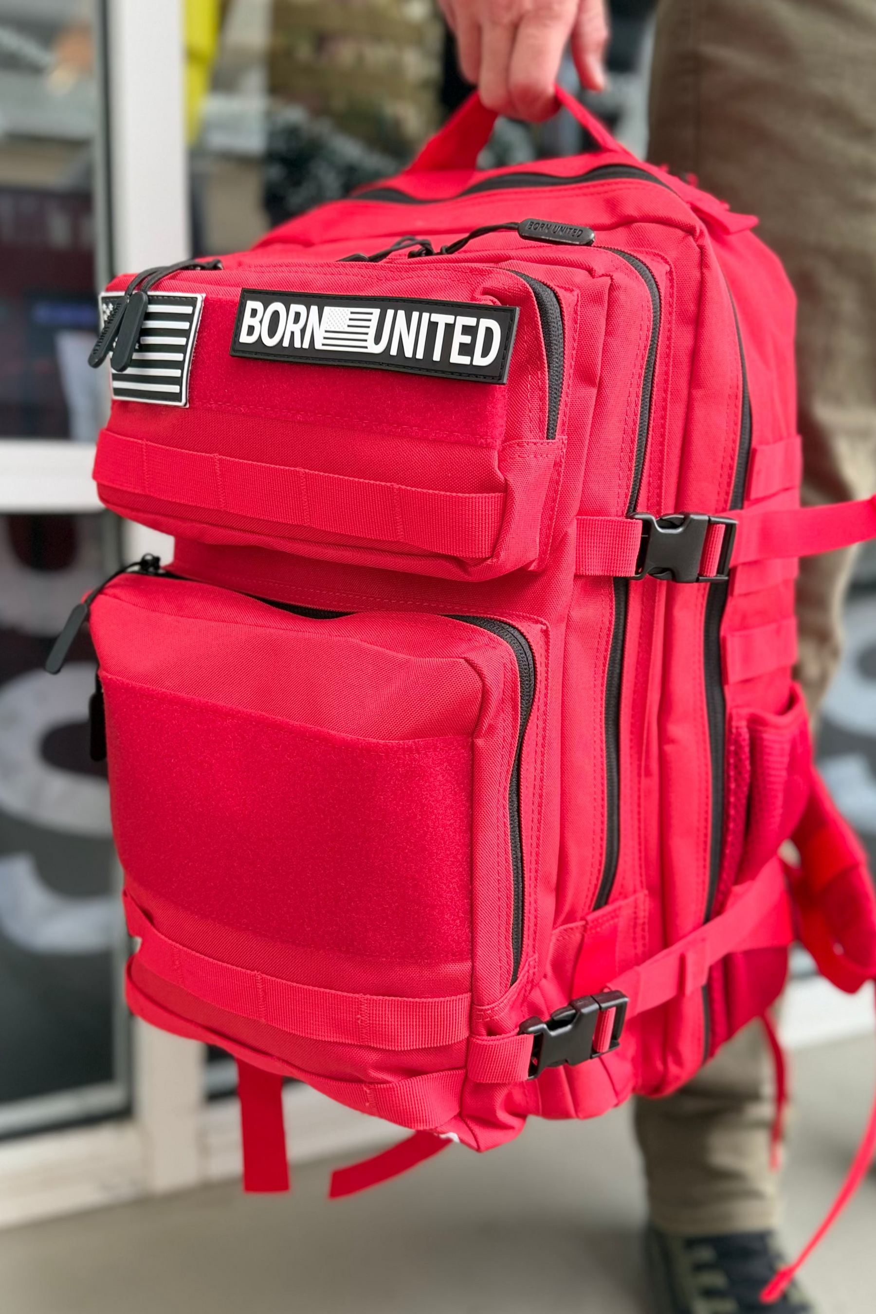 Red backpack with 'BORN UNITED' logo held by a person in an indoor setting.