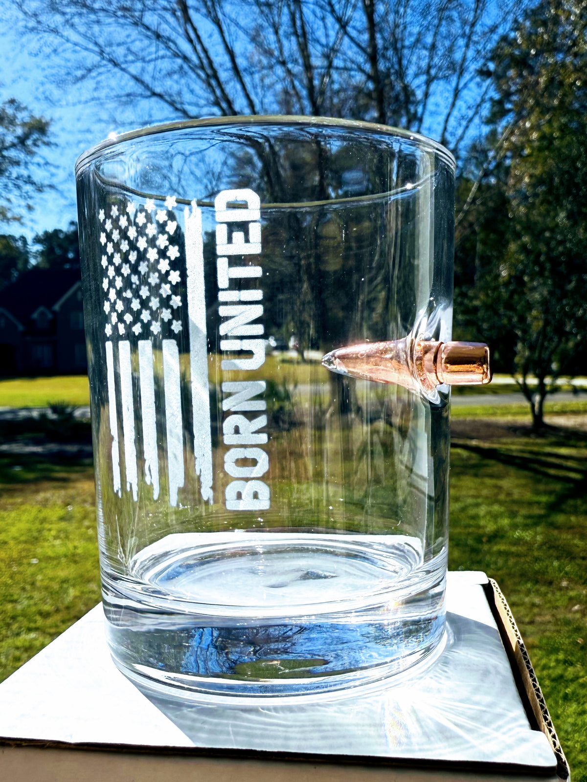 The Born United Bullet Whiskey Glass by Born United Private Label features a distressed American flag graphic with "BORN UNITED" etched on clear glass, plus a copper bullet embedded in the side.