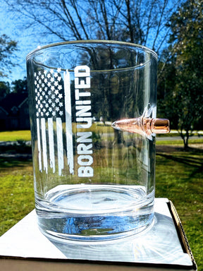 The Born United Bullet Whiskey Glass by Born United Private Label features a distressed American flag graphic with "BORN UNITED" etched on clear glass, plus a copper bullet embedded in the side.