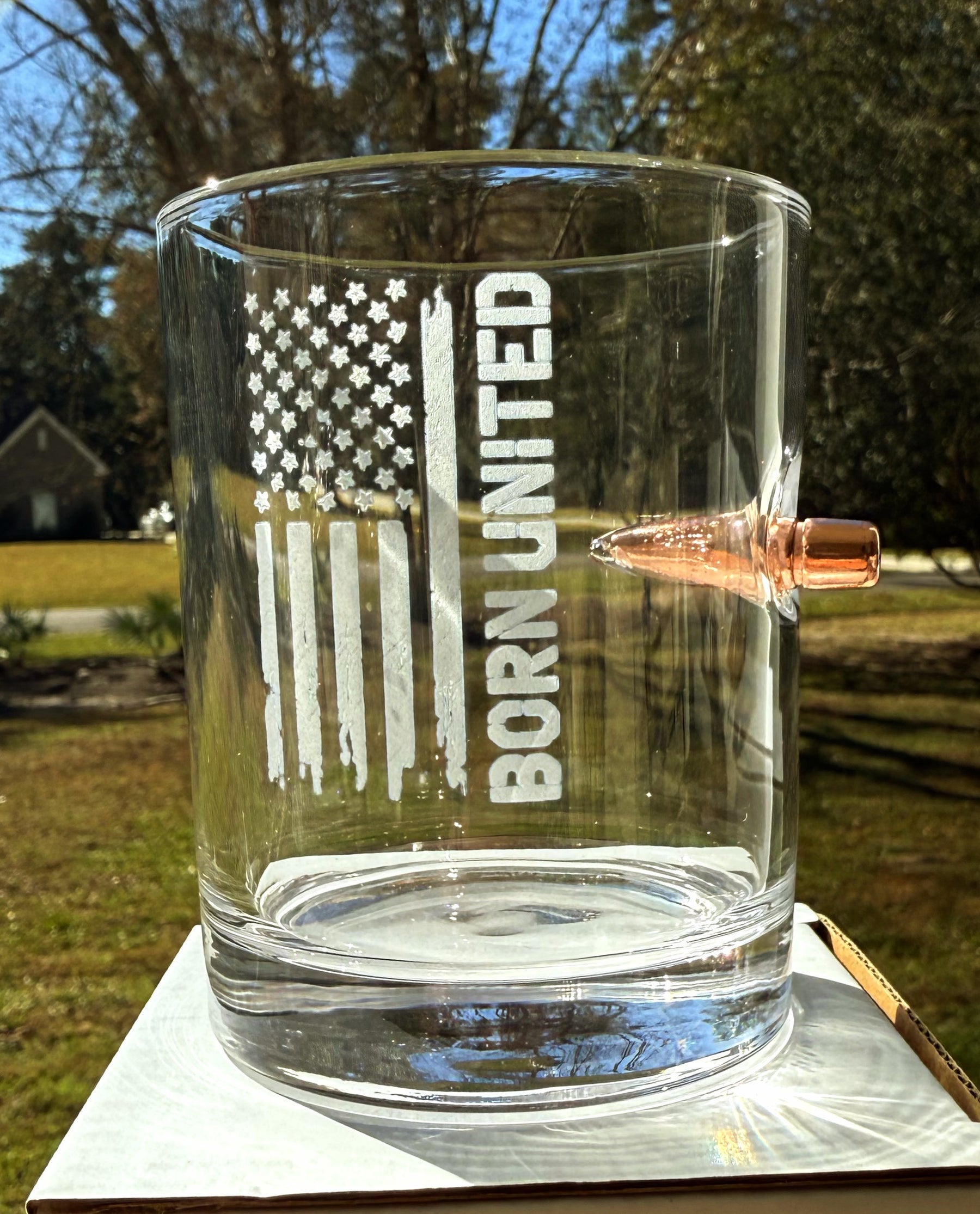 The Born United Bullet Whiskey Glass by Born United Private Label features a real bullet embedded in the side and is etched with a distressed American flag and "BORN UNITED," shown outdoors among grass, trees, and houses.