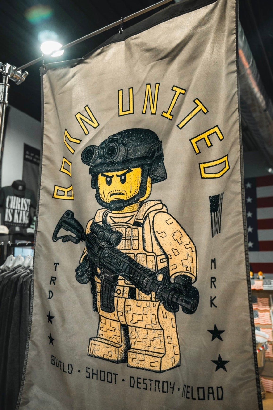The Brick Operator Flag by Born United Private Label displays a Lego-style soldier with a rifle indoors. "BORN UNITED" appears above, with "BUILD • SHOOT • DESTROY • RELOAD" below, accented by stars and small graphics.