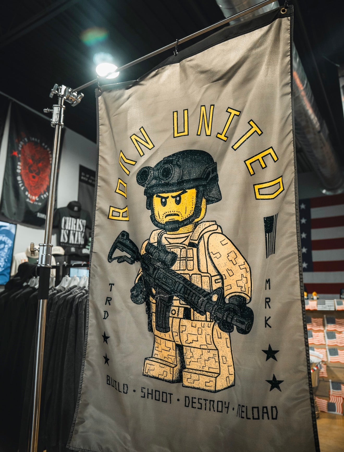 The Brick Operator Flag by Born United Private Label displays a Lego-style soldier with a rifle indoors. "BORN UNITED" appears above, with "BUILD • SHOOT • DESTROY • RELOAD" below, accented by stars and small graphics.