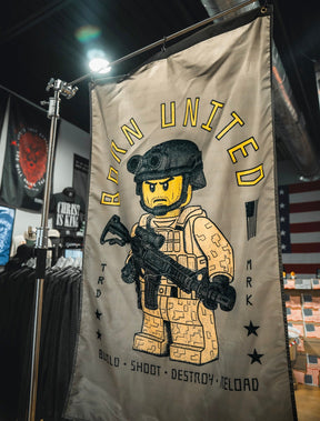 The Brick Operator Flag by Born United Private Label displays a Lego-style soldier with a rifle indoors. "BORN UNITED" appears above, with "BUILD • SHOOT • DESTROY • RELOAD" below, accented by stars and small graphics.