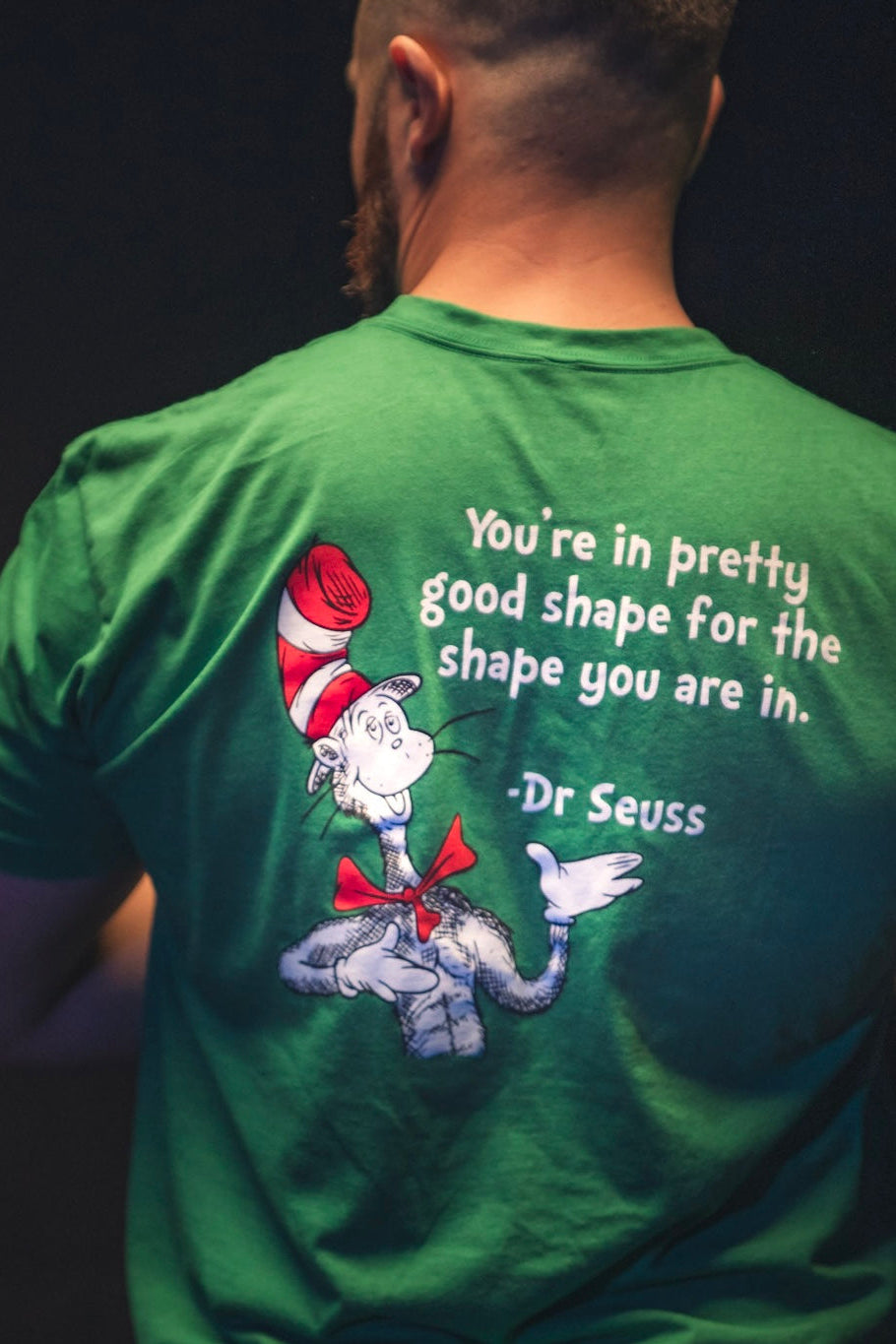 A person wearing the St. Seuss Tee by Tango Charlie Apparel—a green workout shirt with the Cat in the Hat and Dr. Seuss's quote, "You’re in pretty good shape for the shape you are in." Ideal gym motivation for your fitness journey.