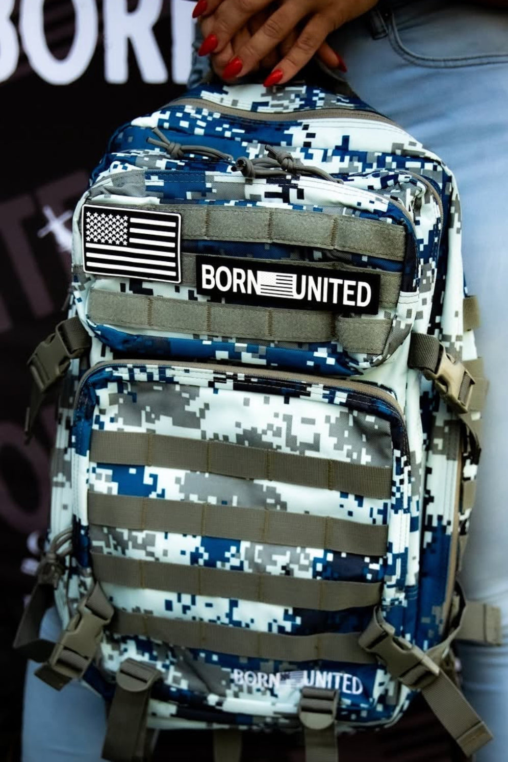 Someone carries the Arctic Tac-Pac - 45L camo backpack by Born United Private Label, featuring an American flag and Born United patch for an adventurous Arctic vibe. They sport a smartwatch and light jeans against a backdrop with partially visible text.