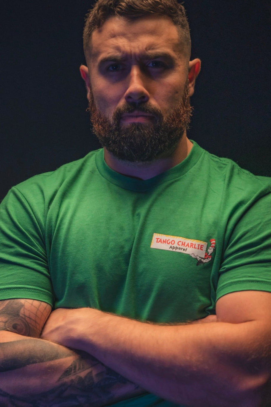 A bearded man with tattoos crosses his arms and looks serious, wearing the green St. Seuss Tee by Tango Charlie Apparel against a dark background, embodying true gym motivation.