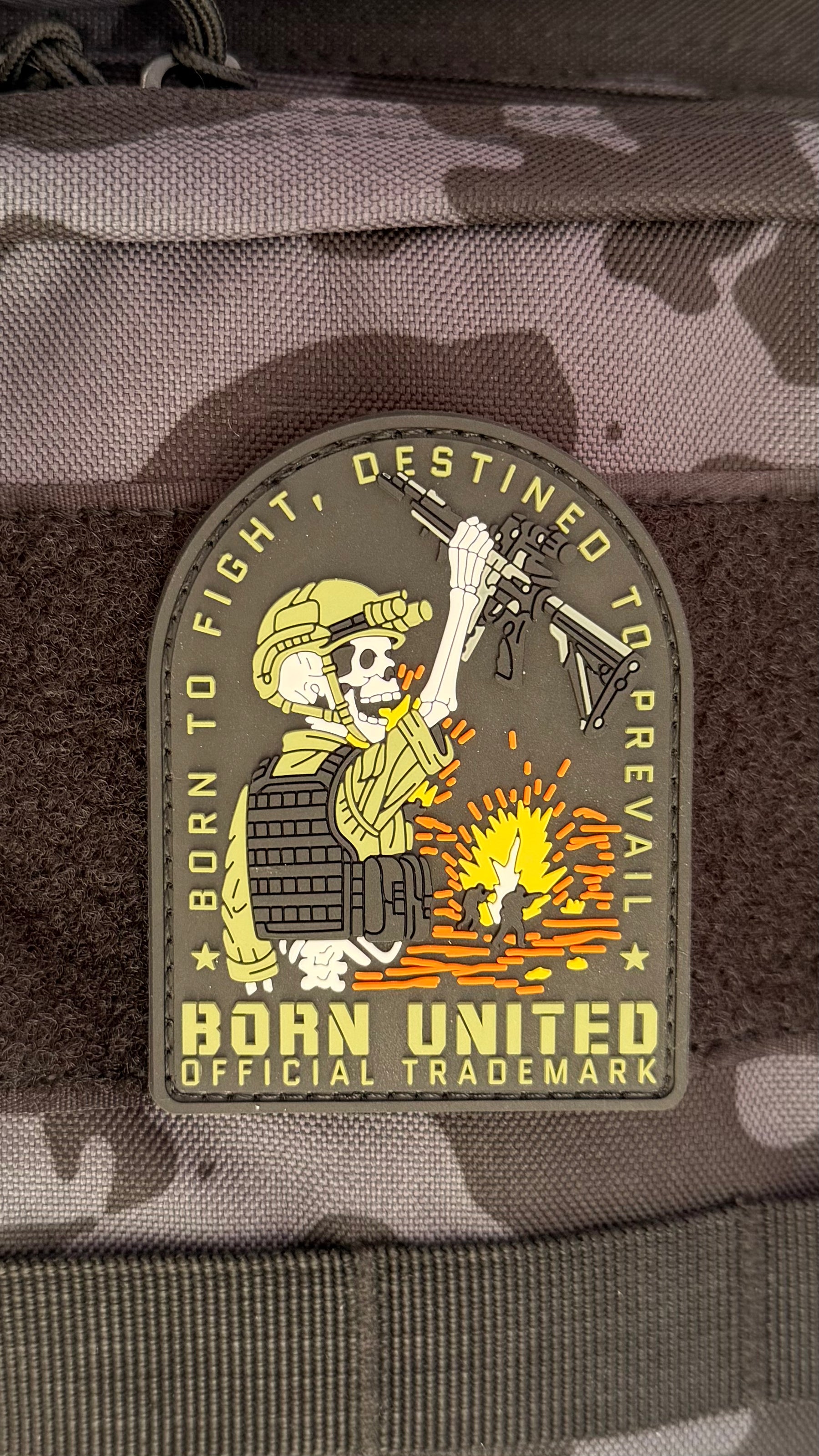 The Destined to Prevail PVC Patch by Born United features a skeleton soldier in tactical gear holding a rifle over a campfire, with “Born to fight, destined to prevail” and “Born United Official Trademark” on a camo background.
