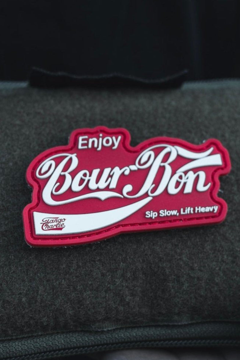 In the dim light, someone holds a zippered pouch with a Tango Charlie Apparel patch saying, Enjoy Bour-Bon, Sip Slow, Lift Heavy, highlighted by its striking red and white colors.