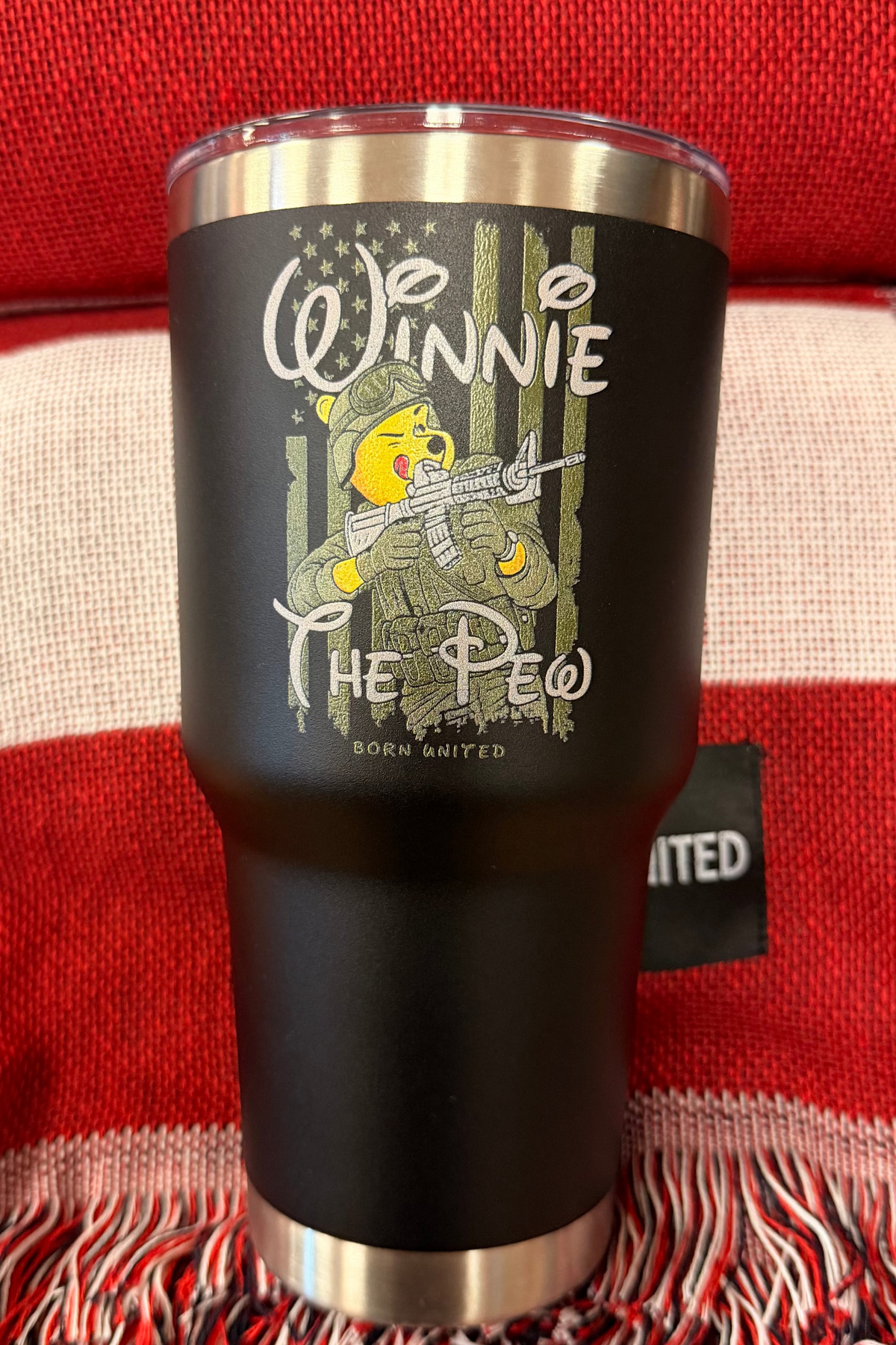 The Winnie the Pew Tumbler - 30oz by Born United Private Label features a bear in army gear with a rifle, "Winnie The Pew" text, and an American flag background on black stainless steel, shown on a red and white woven blanket.