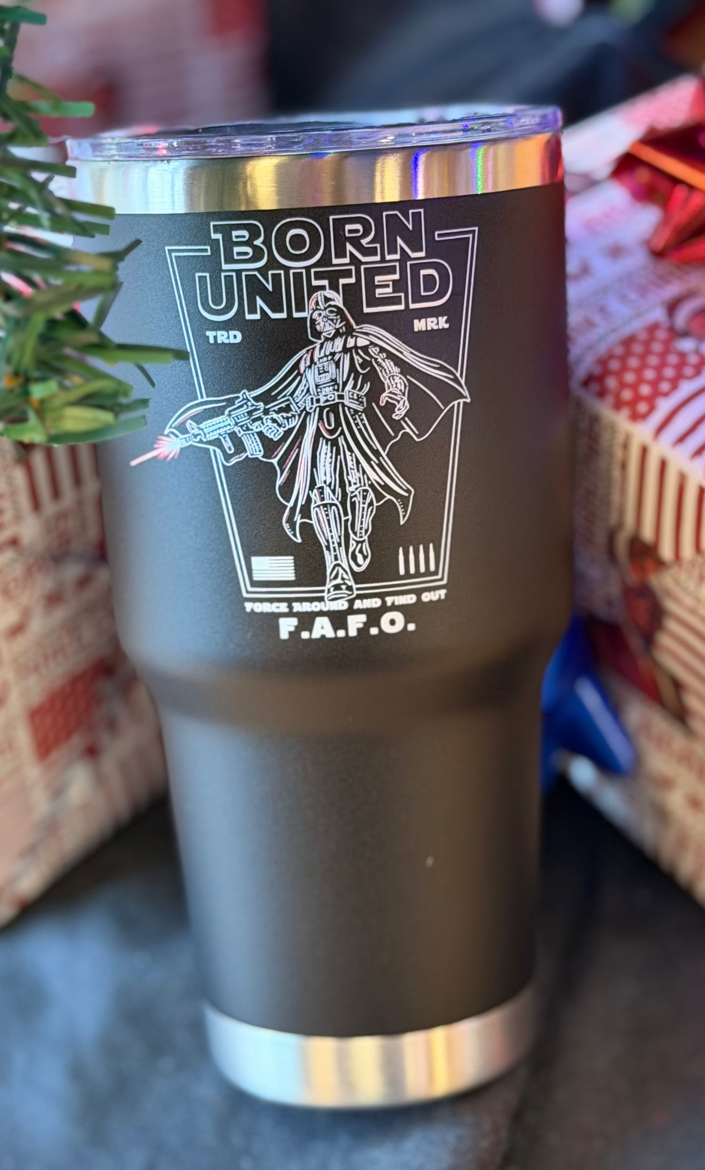 The Born United Private Label Vader FAFO Tumbler - 30oz is a black insulated tumbler featuring a white futuristic soldier graphic and BORN UNITED F.A.F.O. text, shown on a surface near a decorated tree and wrapped gifts.