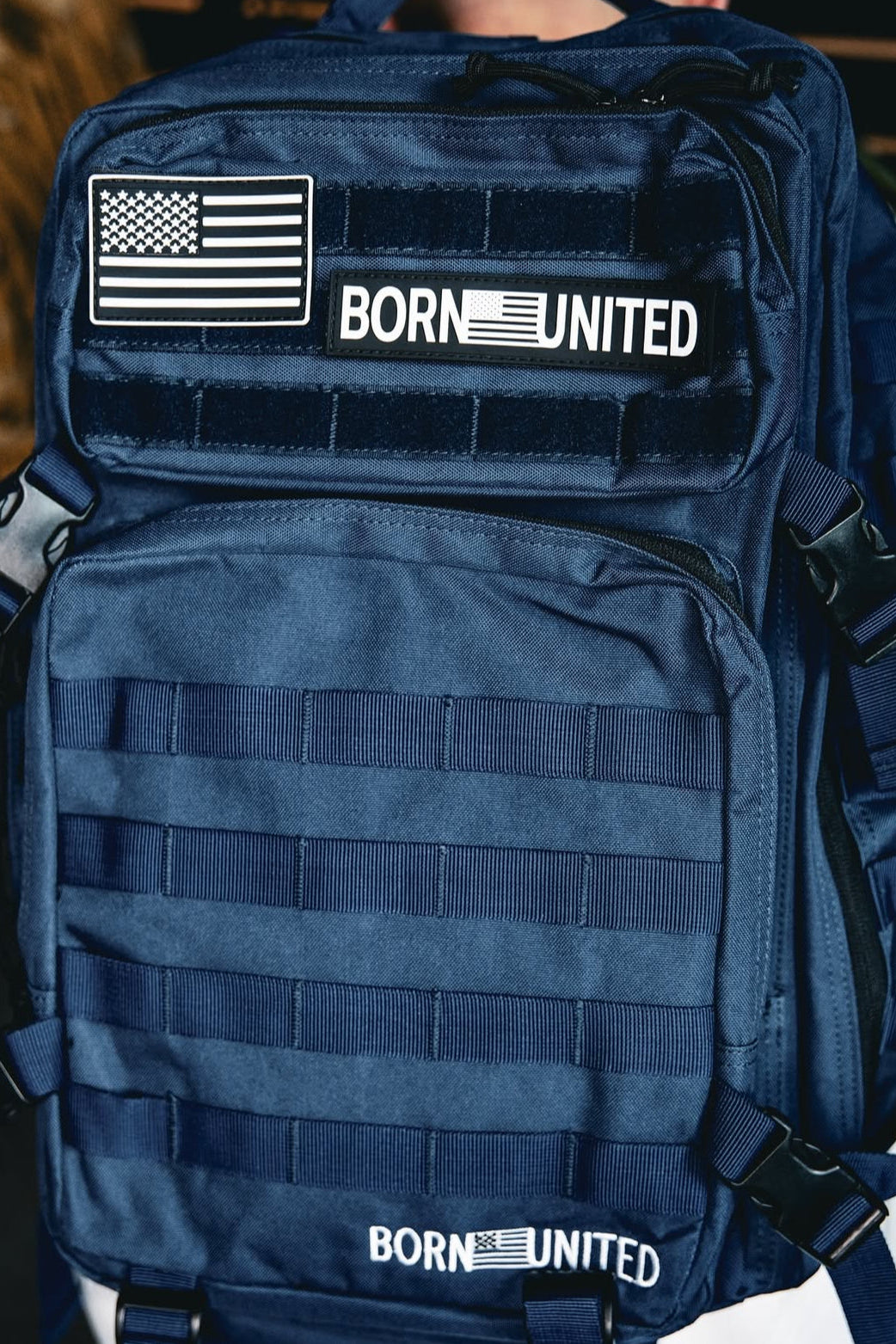 The Navy Blue Tac-Pac - 45L by Born United Private Label showcases the BORN UNITED logo and an American flag patch on the front. With multiple straps and zippers, this durable, functional design is ideal for outdoor or military use.