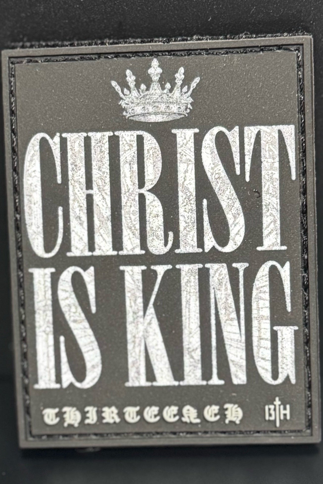 The Christ is King PVC Patch by 13th features a crown above bold white CHRIST IS KING text, ornate THIRTEEN at the bottom, and bH initials on the right, all on a dark background.