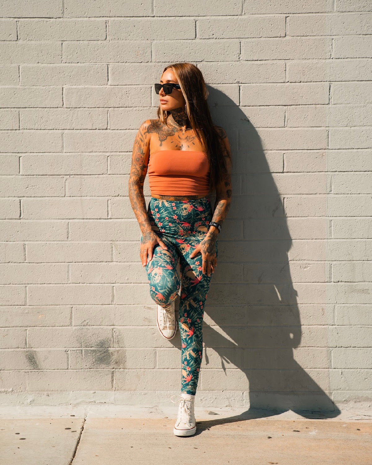 A woman with long hair and tattoos poses against a light brick wall, wearing sunglasses, an orange top, white sneakers, and Tango Charlie Apparel's Women's Performance Leggings | Aloha AR with a floral print. One leg is bent as she looks to the side.