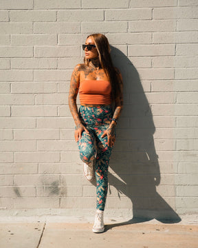 A woman with long hair and tattoos poses against a light brick wall, wearing sunglasses, an orange top, white sneakers, and Tango Charlie Apparel's Women's Performance Leggings | Aloha AR with a floral print. One leg is bent as she looks to the side.