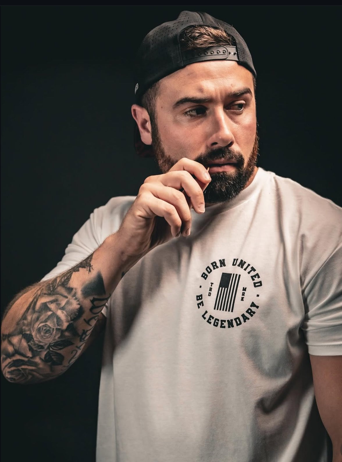 A bearded man in a white Legends - Andy Griffith T-shirt from Born United Private Label stands against a dark background. Wearing a backward black cap, he displays tattoos on his left arm. The premium tee features a circular design with Born United Be Legendary, reflecting American cultures spirit.