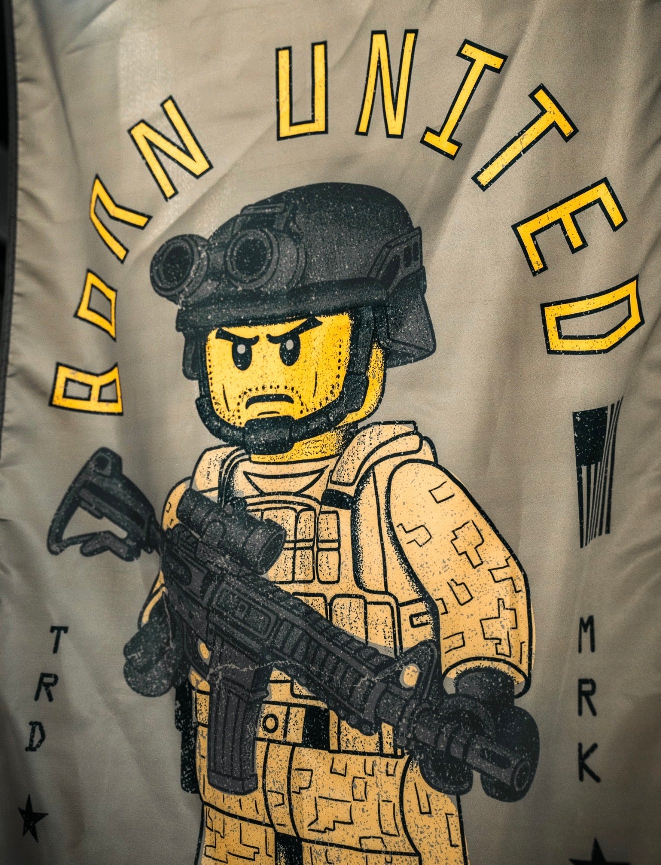 A LEGO-style soldier in desert camo with a rifle and helmet featuring night-vision goggles stands under the bold yellow "Born United" logo and the Brick Operator Flag by Born United Private Label.