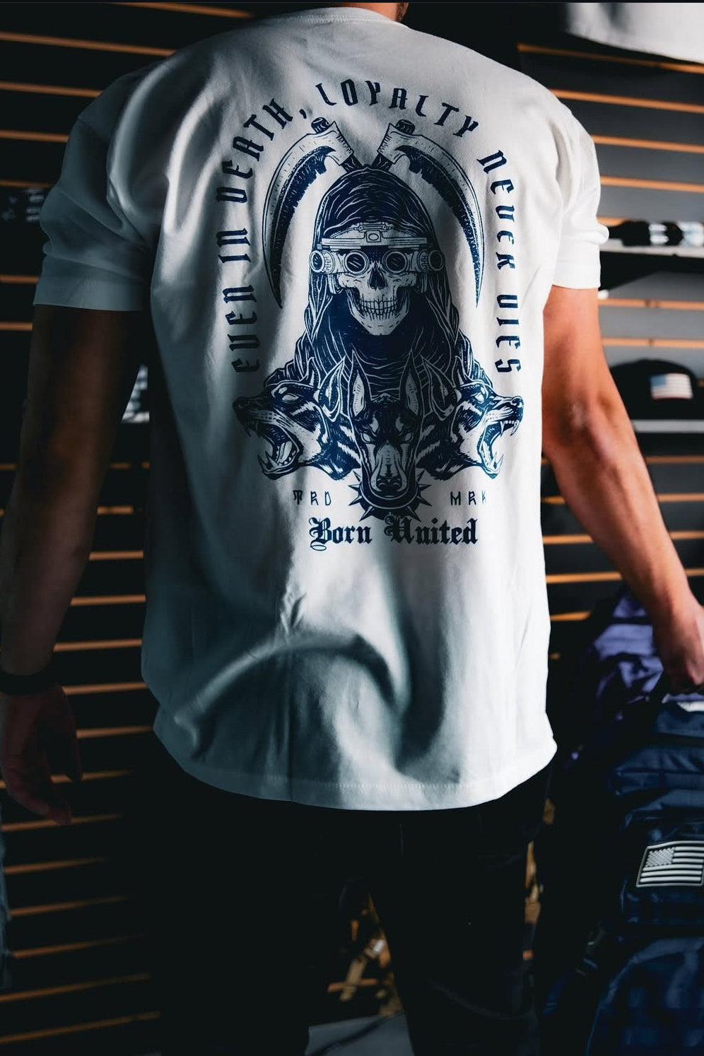 In a store with hats and bags on display, someone wearing a white shirt adorned with a large skull design on the back holds the Navy Blue Tac-Pac - 45L by Born United Private Label.