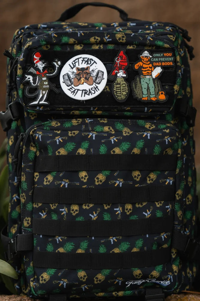 The TCA Tac-Pac Tropic Thunder 2.0 by Born United Private Label is a black backpack with colorful skull and cartoon patches, offering tactical versatility. With its waterproof design, its perfect for outdoor adventures and stands out in lush green surroundings.