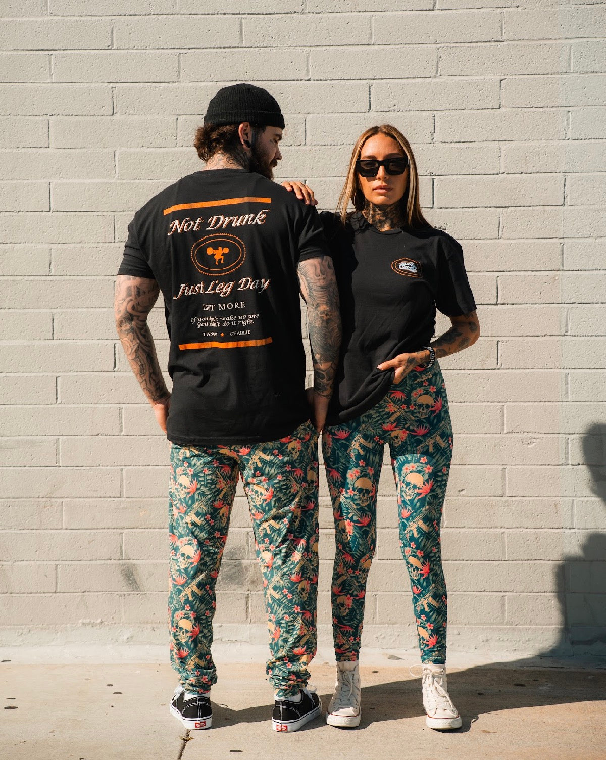 A man and woman pose by a light brick wall in black tees and high rise leggings. The woman, wearing sunglasses and holding her shirt, sports the Women's Performance Leggings | Aloha AR from Tango Charlie Apparel. The man stands facing away.