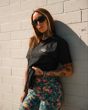 A woman with tattoos wearing sunglasses, a black t-shirt, and Tango Charlie Apparel’s Women's Performance Leggings | Aloha AR stands against a sunlit white brick wall, holding her shirt and looking confidently at the camera.